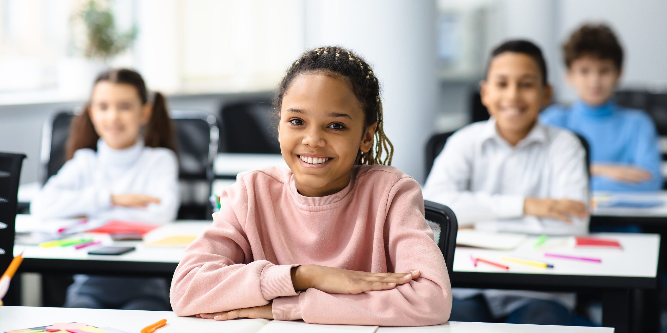 Elementary Education. Portrait of happy small african american girl sitting at table in classroom at primary school with group of classmates and posing, looking at camera and smiling